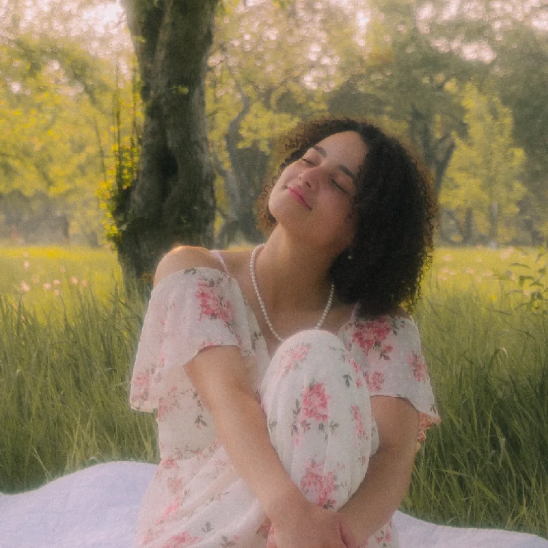 Elisha Antoine smiling with eyes closed in a sunny meadow, wearing a floral dress and pearls
